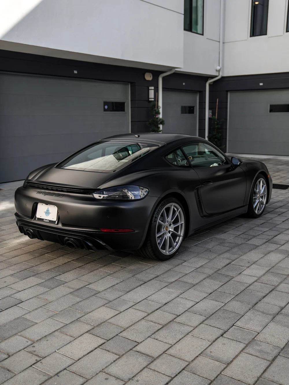 Elevated rear three-quarter view of the Porsche 718 Cayman GTS with 3M Satin Black vinyl wrap, showing the rear window and roof