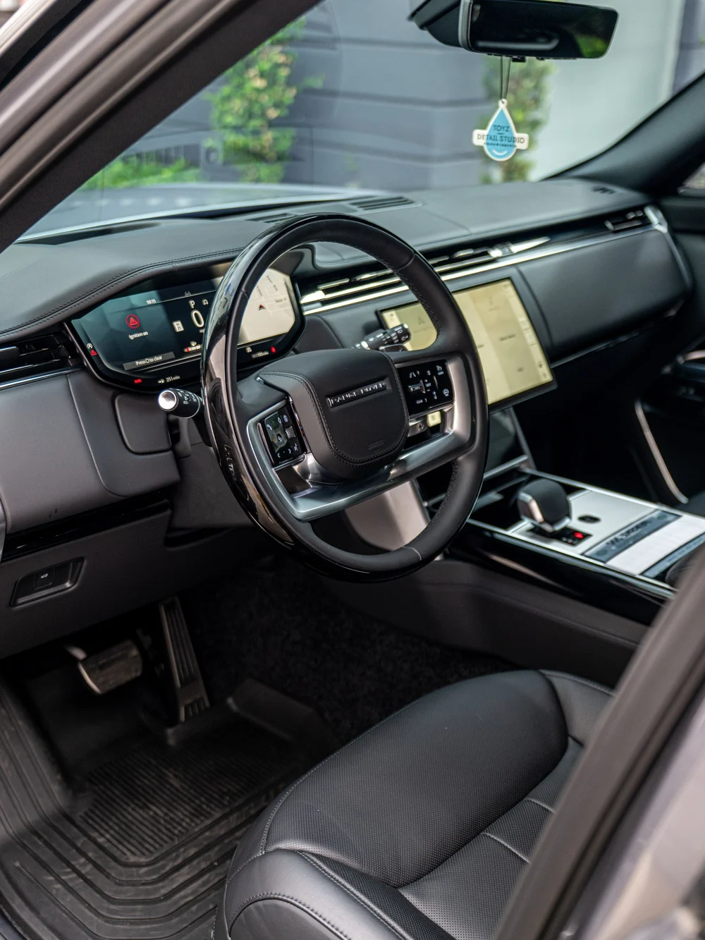 Range Rover driver's cockpit viewed through the open front door, with the steering wheel, dashboard screen and center console visible.