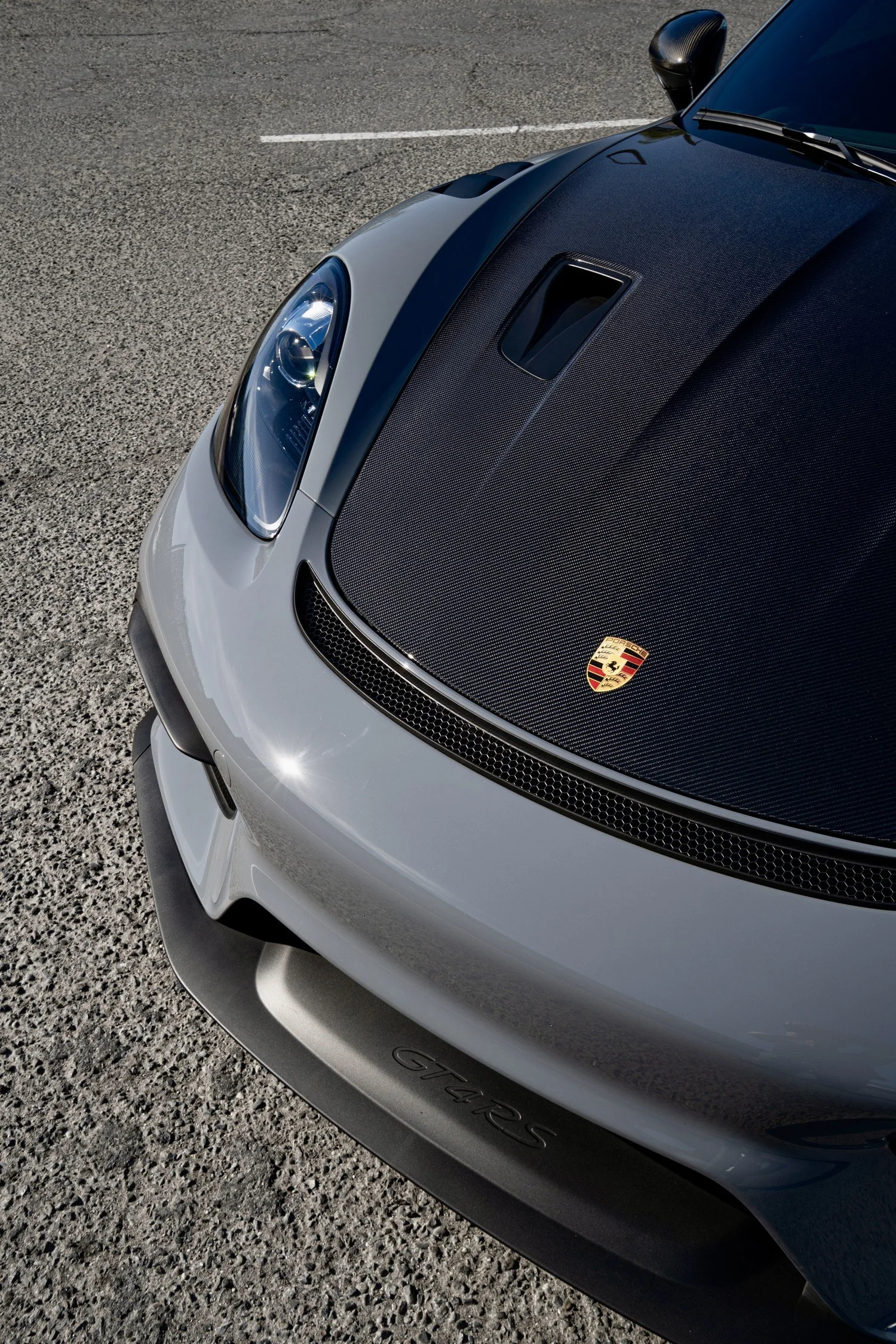 Angled close-up of the GT4 RS carbon hood, Porsche crest, headlight and Arctic Grey front bumper.