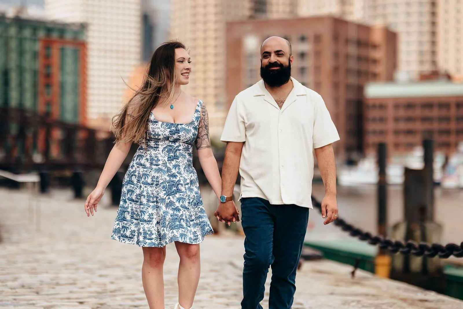 Aziz and Katherine walking hand in hand with the Boston skyline behind them