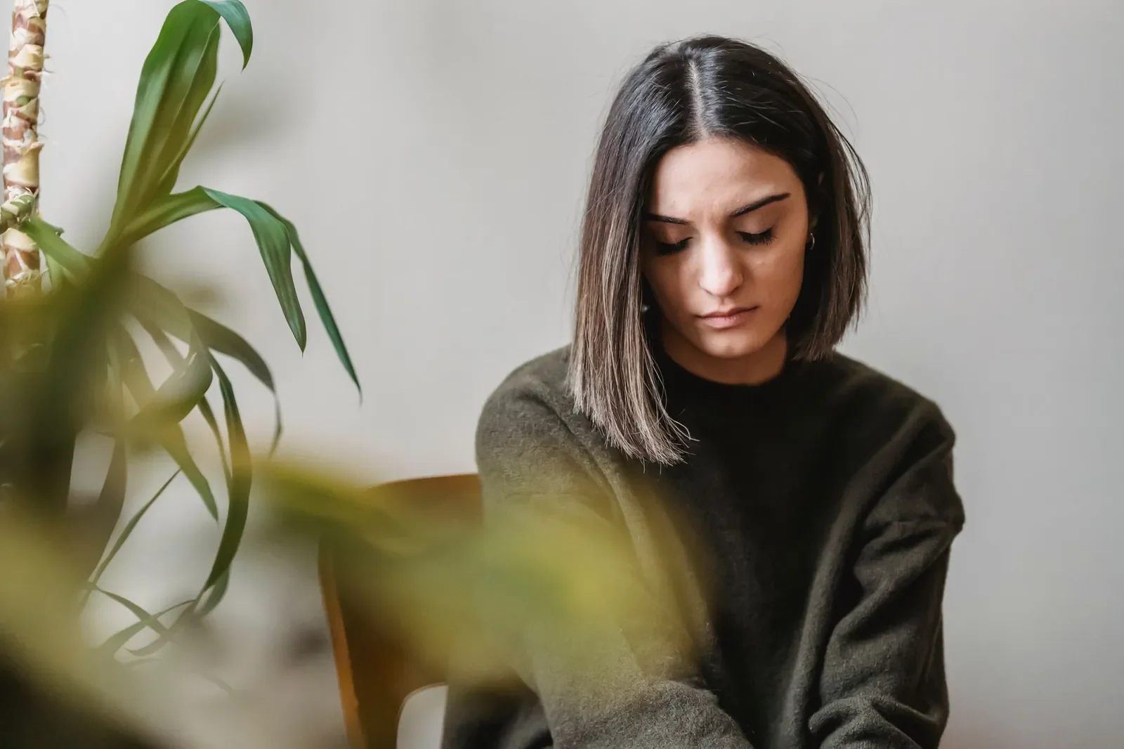 Woman in a dark green sweater sits on a wooden chair looking down beside a plant