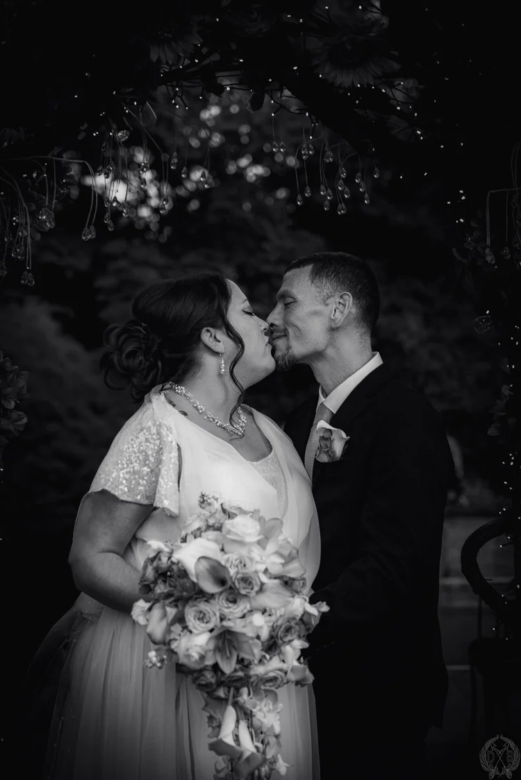 Black-and-white wedding portrait of a bride holding a bouquet and a groom sharing a kiss beneath a floral arch with hanging lights.