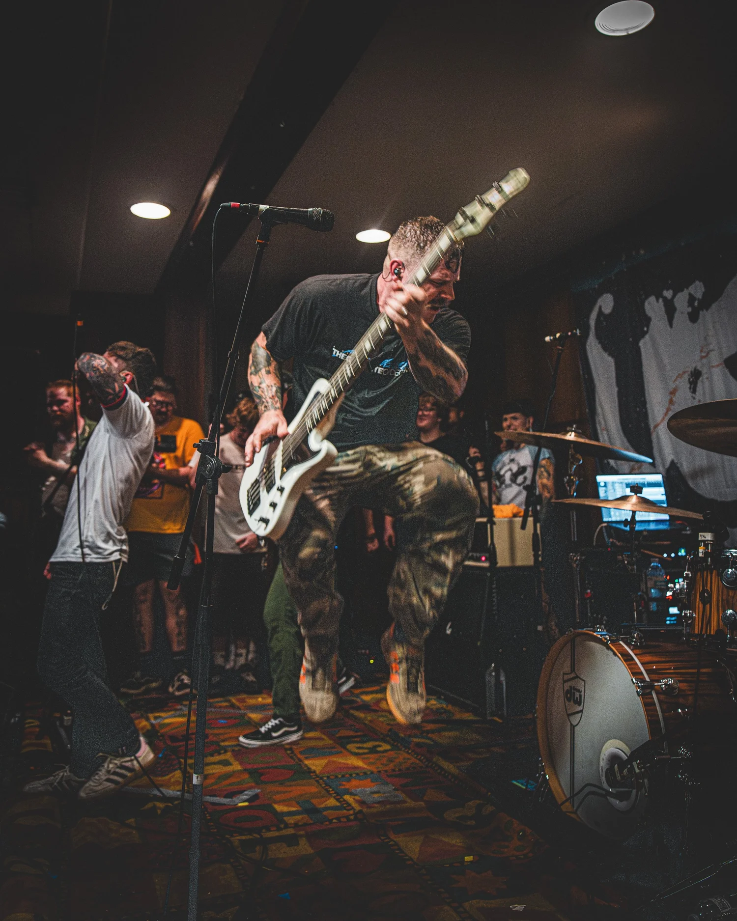 VANNA bassist jumping mid-performance with a white bass as the vocalist and drum kit frame the stage