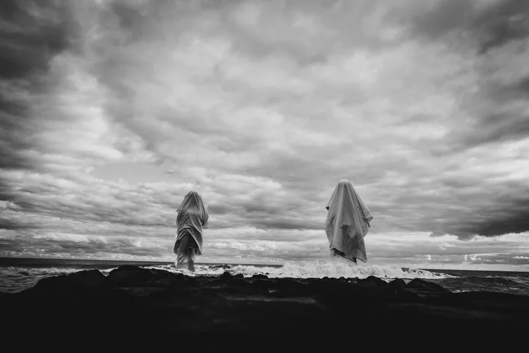 Black-and-white photograph of two draped figures standing on dark rocks beside the ocean beneath dramatic storm clouds.