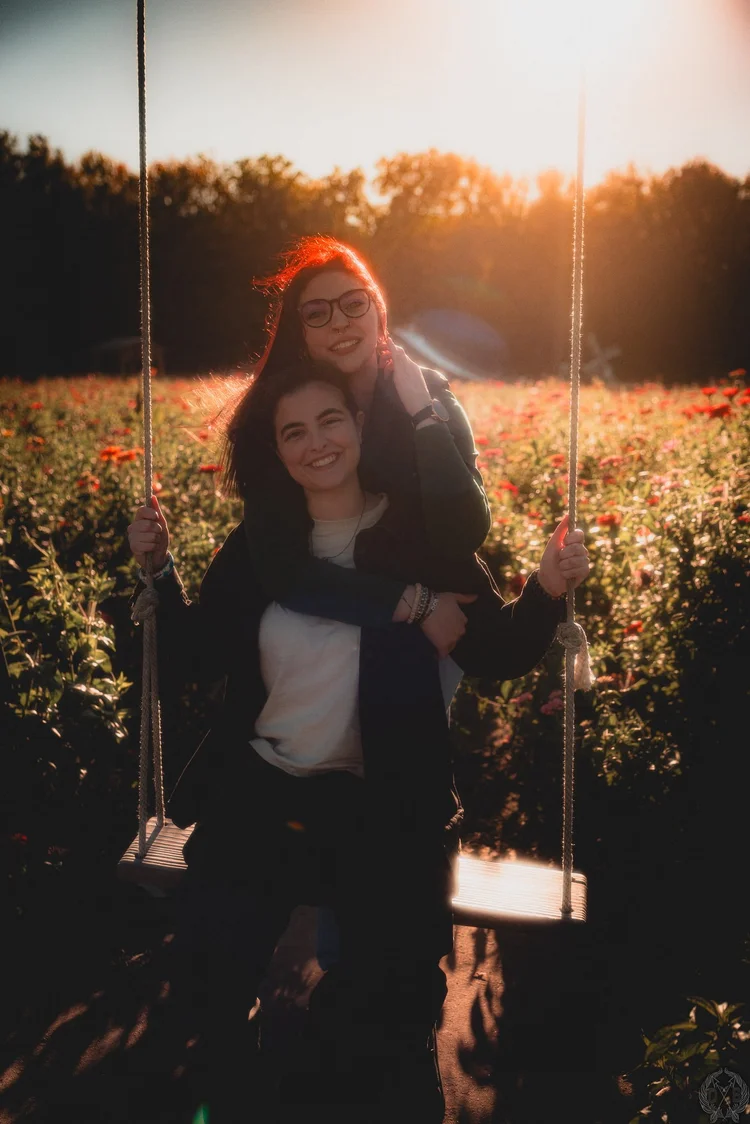 Couple posing together on a swing in a flower field at golden hour, backlit by warm sunlight.