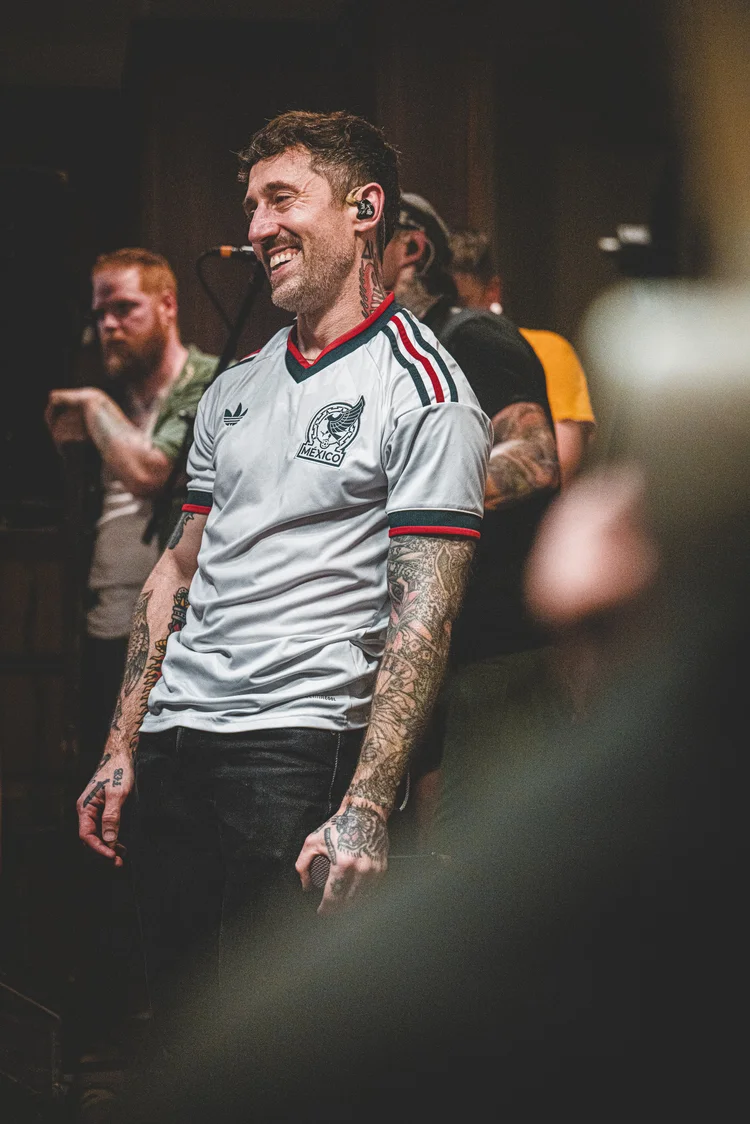 Tattooed musician smiling in a white Mexico jersey during a crowded indoor live show.