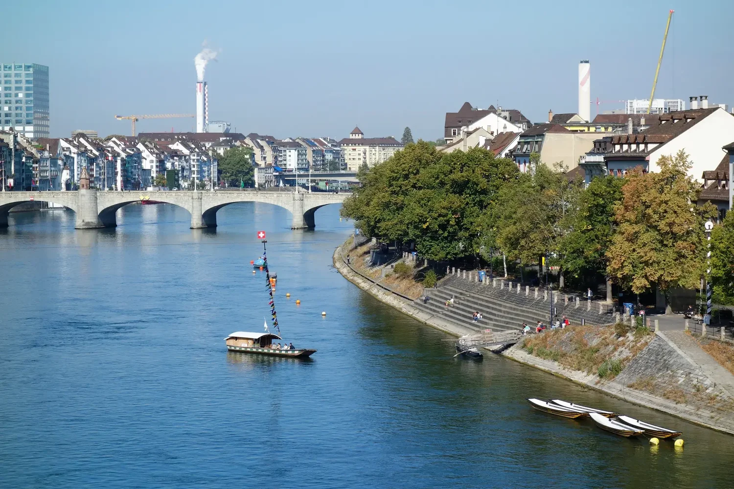 Blick auf den Rhein in Basel mit Steinbrücke, Rheinfähre und Altstadthäusern am Ufer