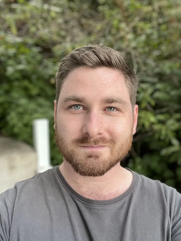 Head and shoulders photo of Callum McMenamin. He has short brown hair, a beard, and is wearing a grey T-shirt, and is looking at the camera.
