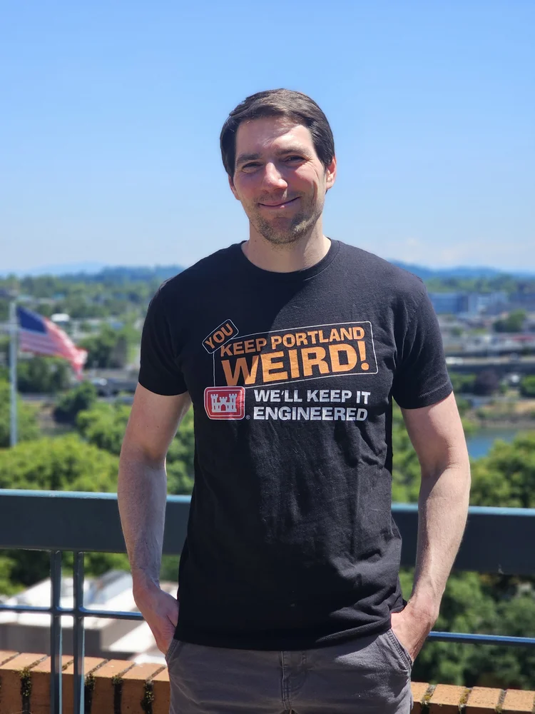 Head and shoulders photo of Callum McMenamin. He has short brown hair, a beard, and is wearing a grey T-shirt, and is looking at the camera.