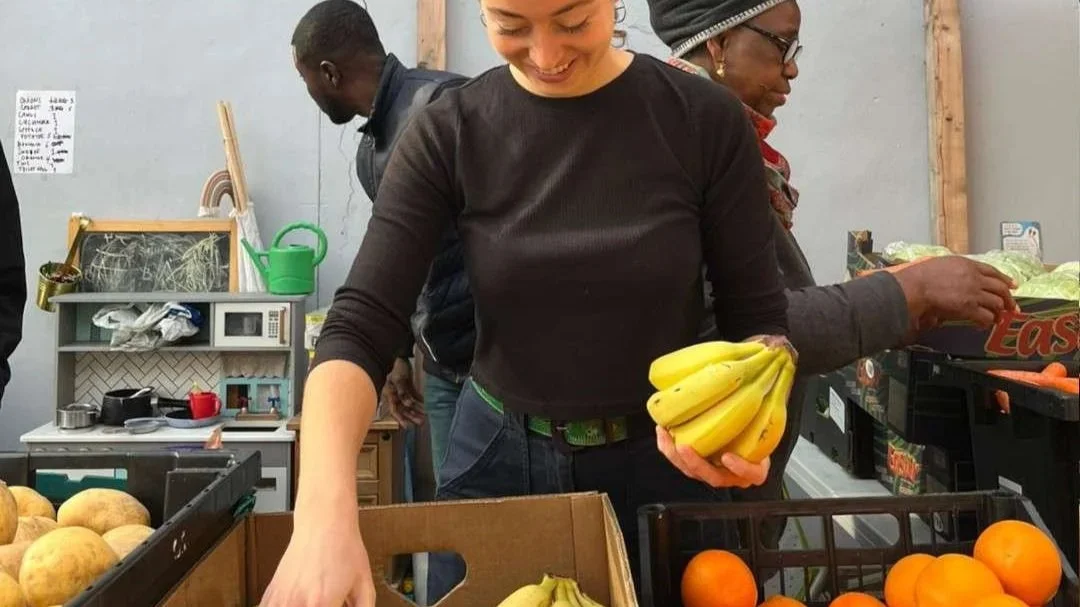 A woman selects bananas with other people walking nearby. The table has bananas, oranges and potatoes. Food & Solidarity sign is visible in the corner.