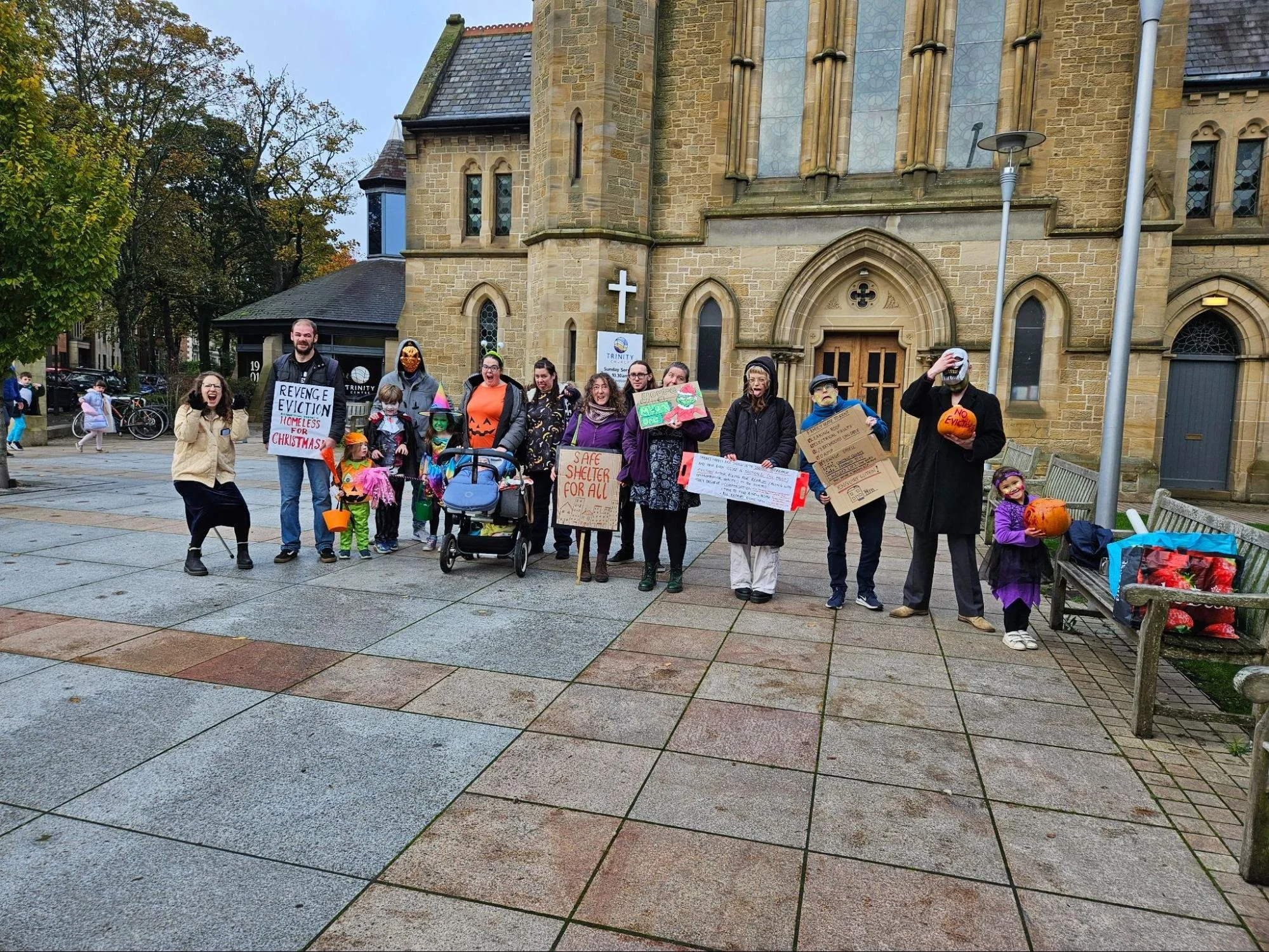 Community Action, food and solidarity members stand in a line in Halloween costumes, placards read 'no evictions' and 'revenge eviction, homeless for christmas'