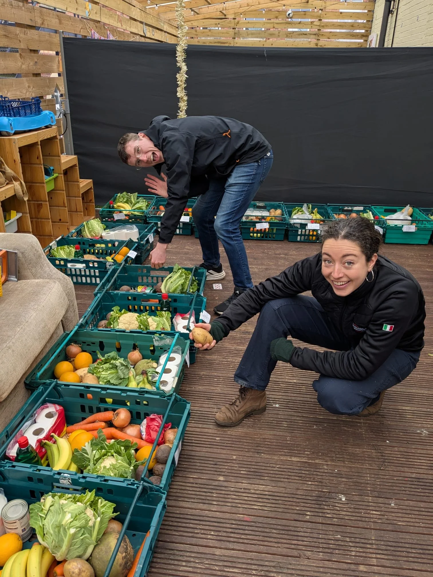 Two Food & Solidarity members smiling and crouching over a row of green crates packed with fresh food parcels including cauliflower, bananas, carrots, oranges, and toilet rolls, ready for Saturday distribution in Newcastle