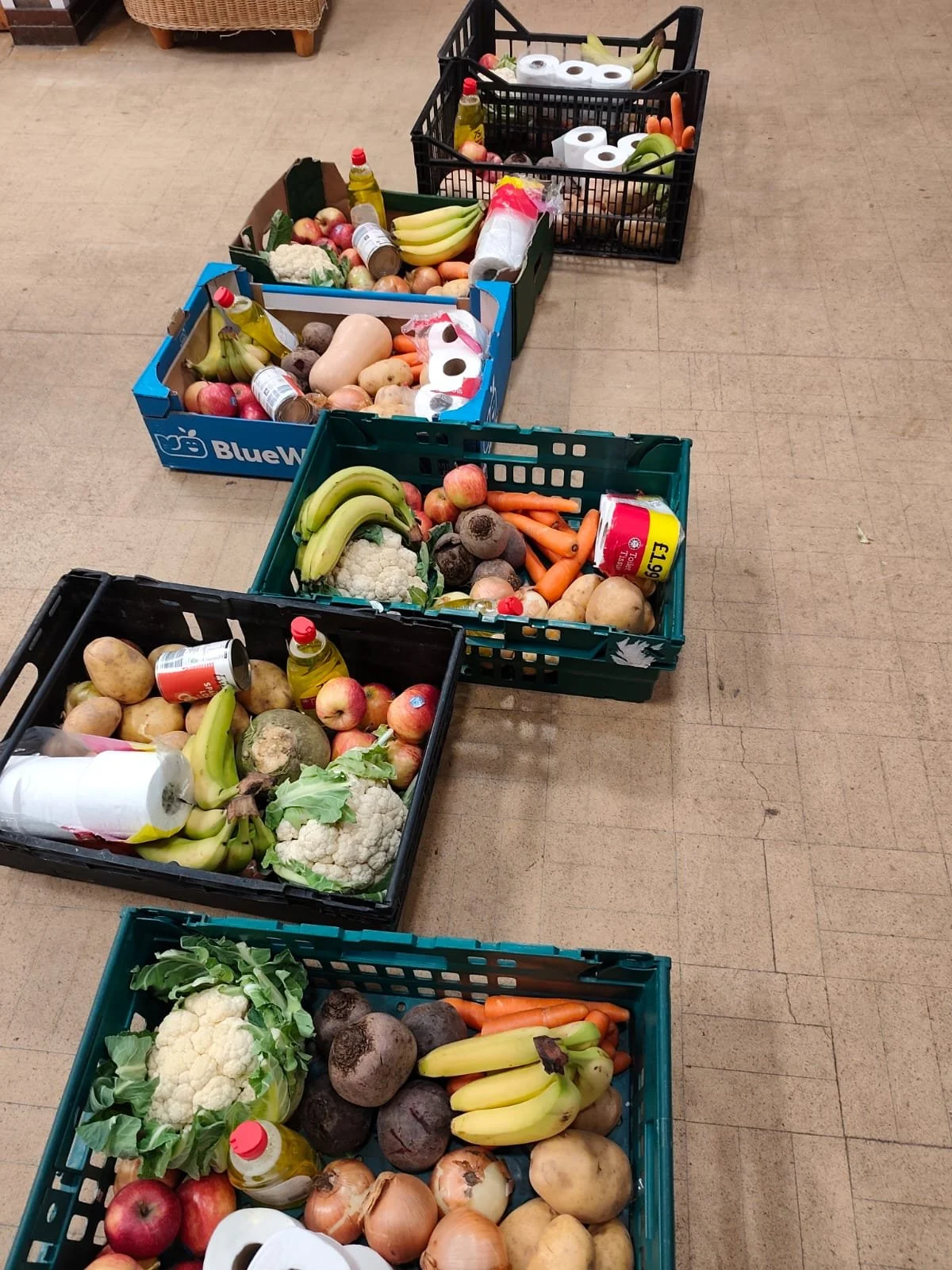 A row of five green crates on a hall floor, each packed with a weekly food parcel containing cauliflower, potatoes, onions, bananas, apples, carrots, cooking oil, tinned goods, and toilet rolls, ready for collection at Food & Solidarity Newcastle, March 2026