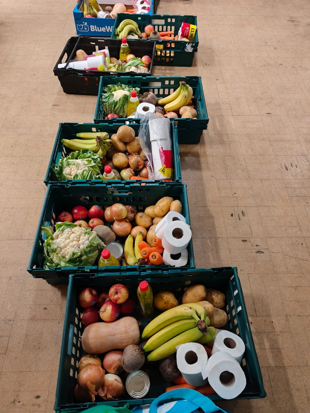 A row of five green crates on a hall floor, each packed with a weekly food parcel containing cauliflower, potatoes, onions, bananas, apples, carrots, cooking oil, tinned goods, and toilet rolls, ready for collection at Food & Solidarity Newcastle, March 2026
