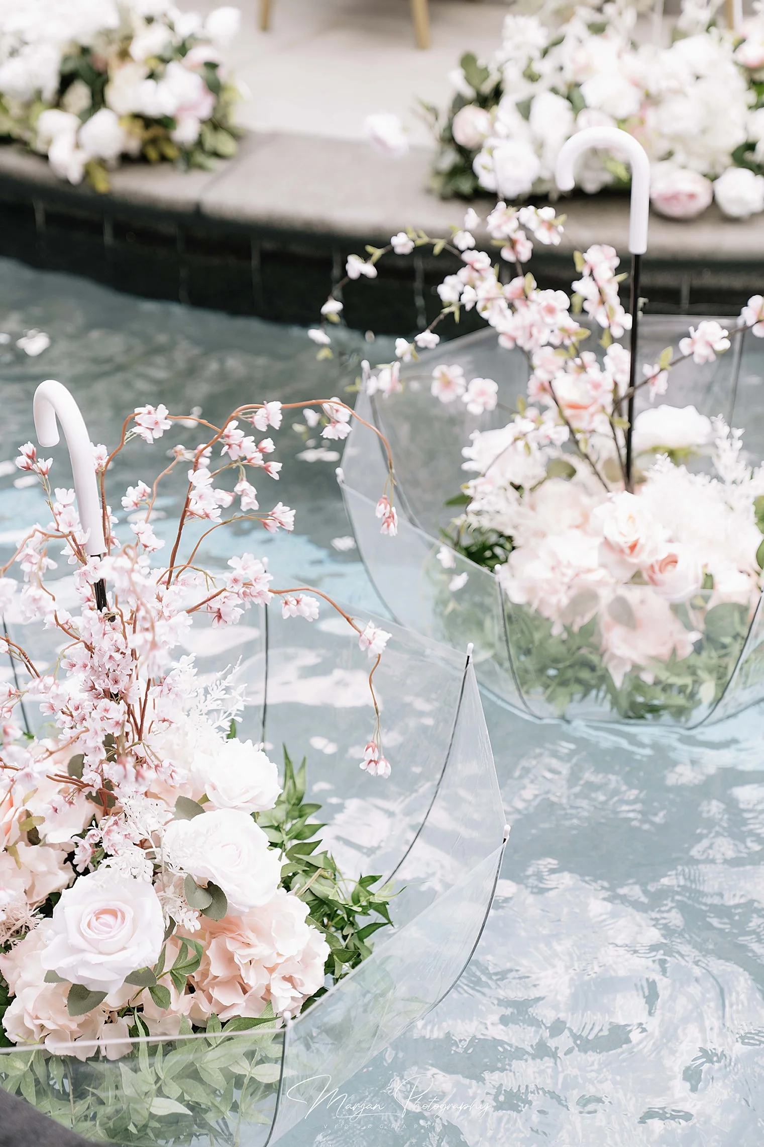 Whimsical poolside engagement decor featuring blush floral arrangements, delicate pink blossoms and clear decorative umbrellas.