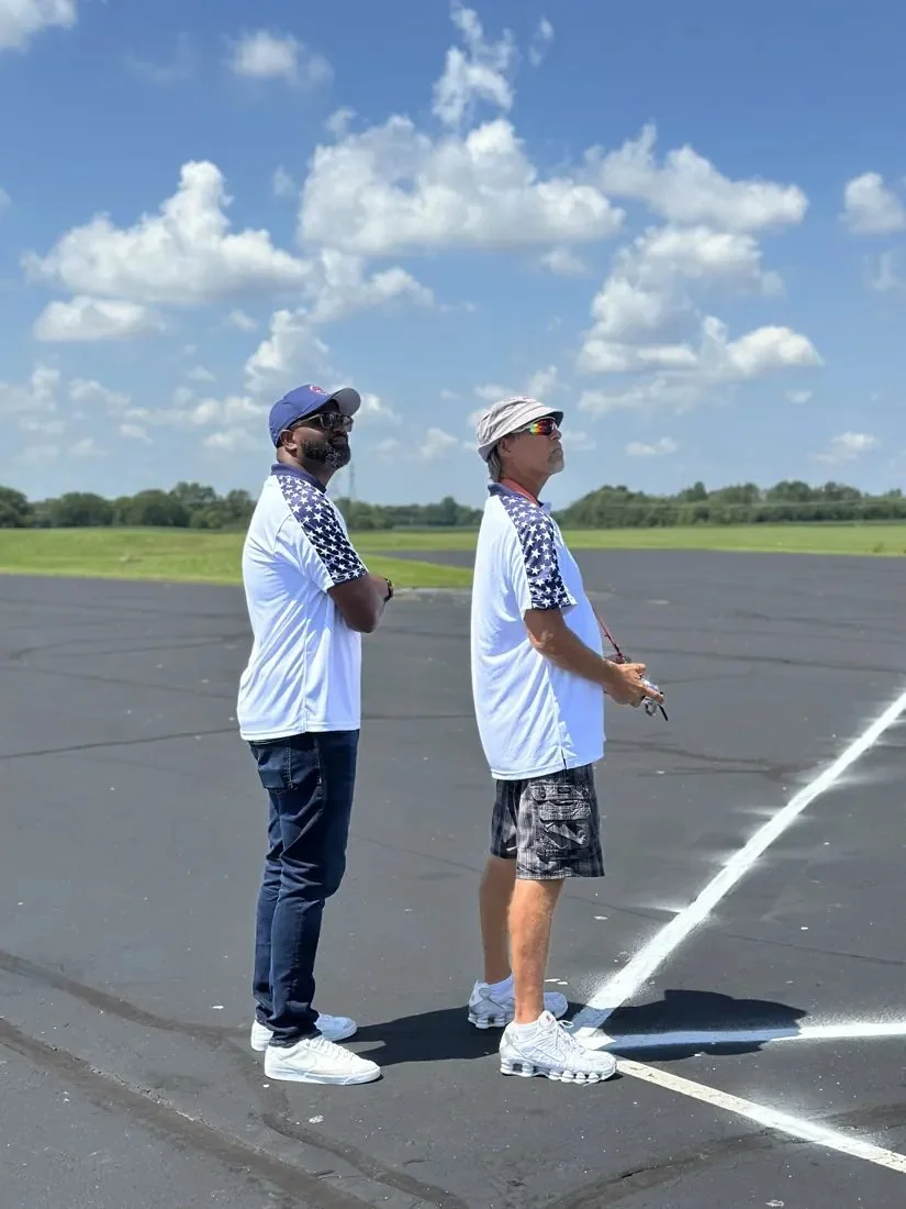 Two Team USA members in stars-and-stripes polos stand side by side on the flightline; the pilot flies with his transmitter while the caller beside him watches the airplane.