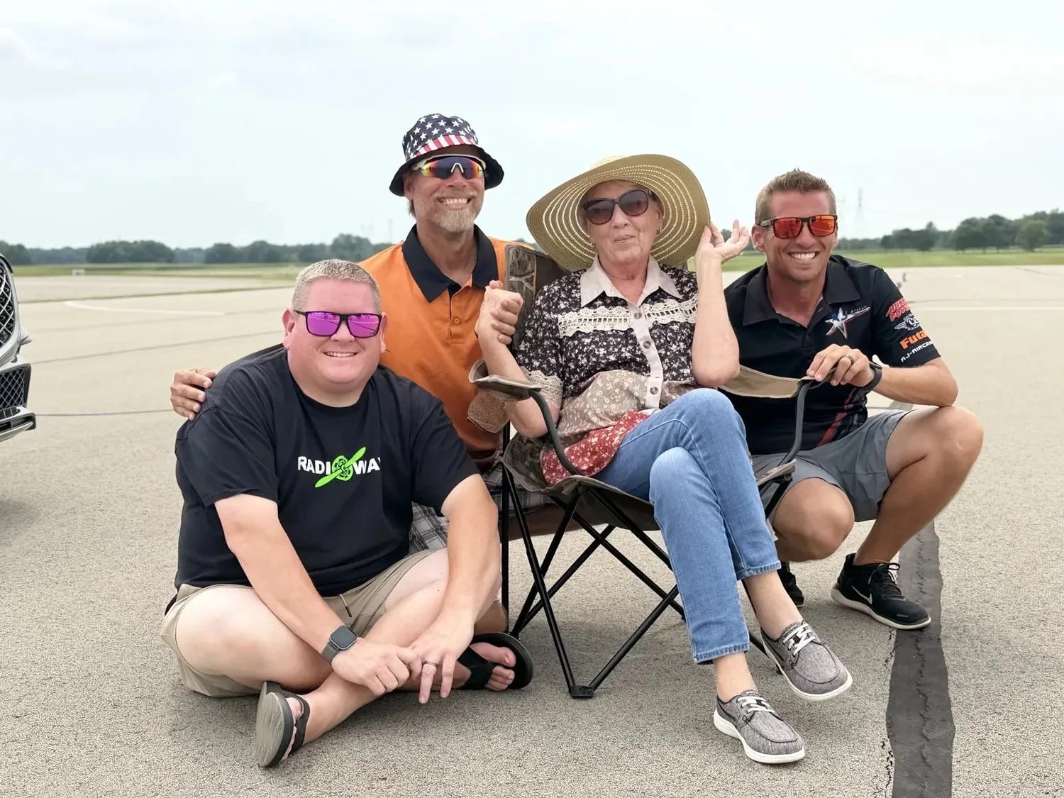 Linda Jesky, in a wide-brimmed sun hat, seated in a folding chair on the flightline surrounded by smiling Team USA members.