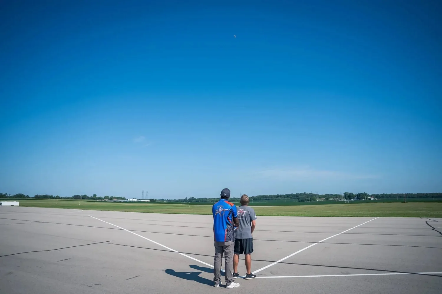 A pilot and caller stand at the flight station on a wide runway under a deep blue sky; the F3A aircraft is a small speck high above them.