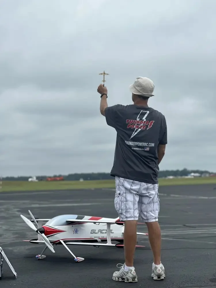 A pilot on the flightline holds a small stick model overhead to rehearse a sequence, with his F3A aircraft on the runway beside him.
