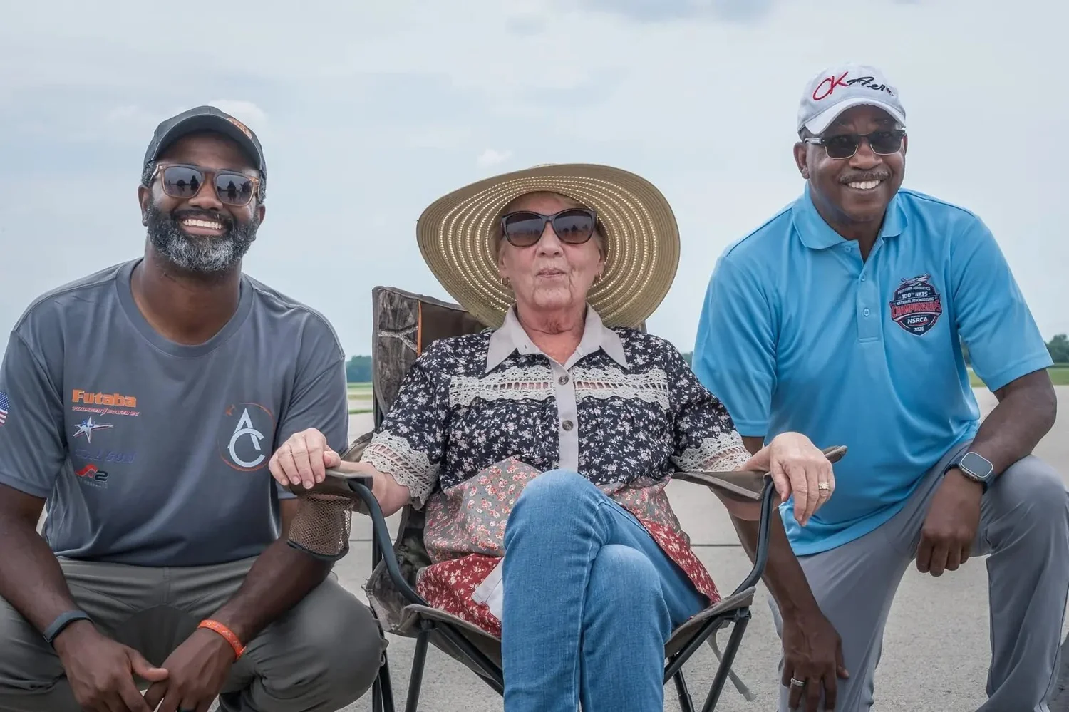 Linda Jesky in a wide straw sun hat, seated in a folding chair on the flightline between AC Glenn and a smiling friend.