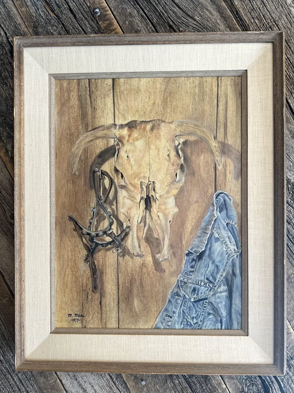 A vintage framed painting of a cow skull, rope and a denim jacket on barn wood