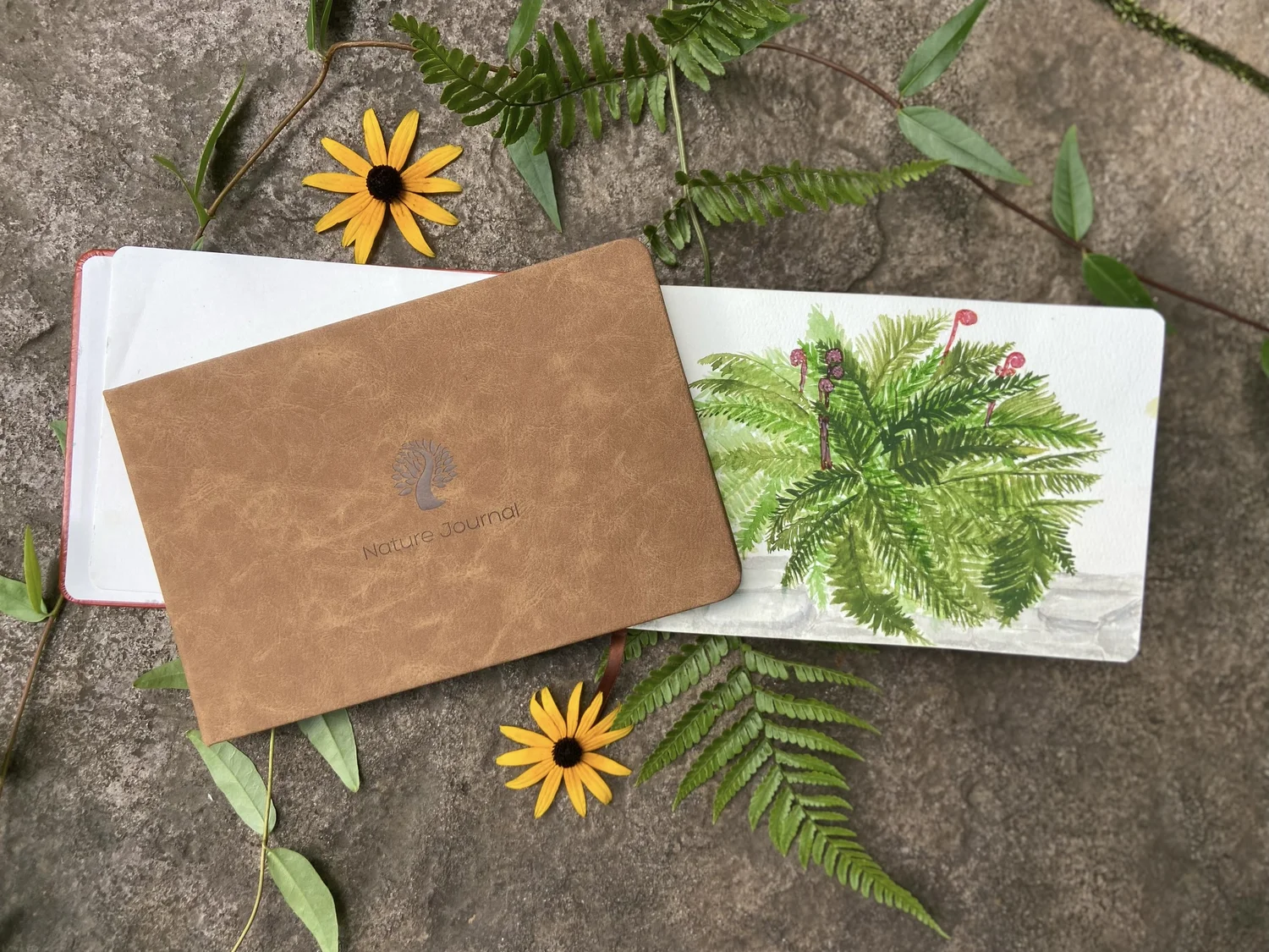 A camel nature journal resting on an open journal painted with a fern, among ferns and black-eyed Susans on stone