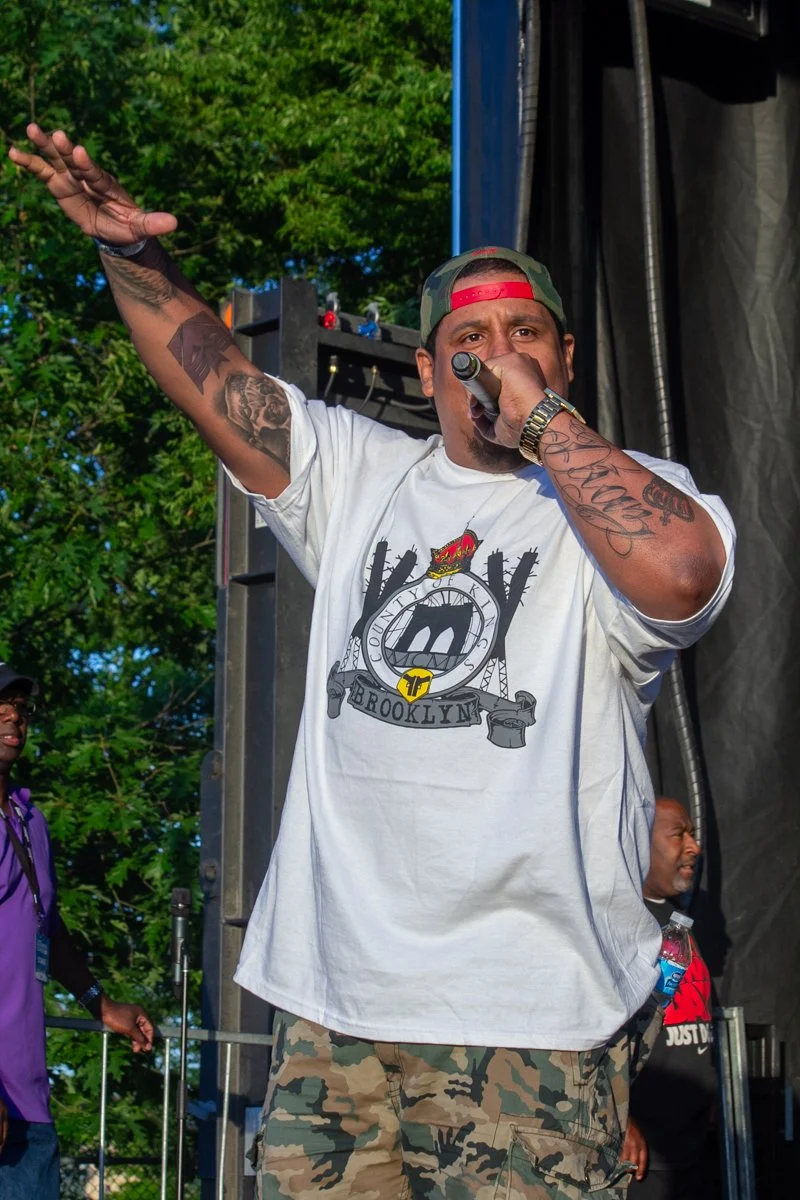 Pumpkinhead performs onstage with a microphone during an outdoor hip-hop concert, joined by fellow MC LR Blitzkrieg and other performers.