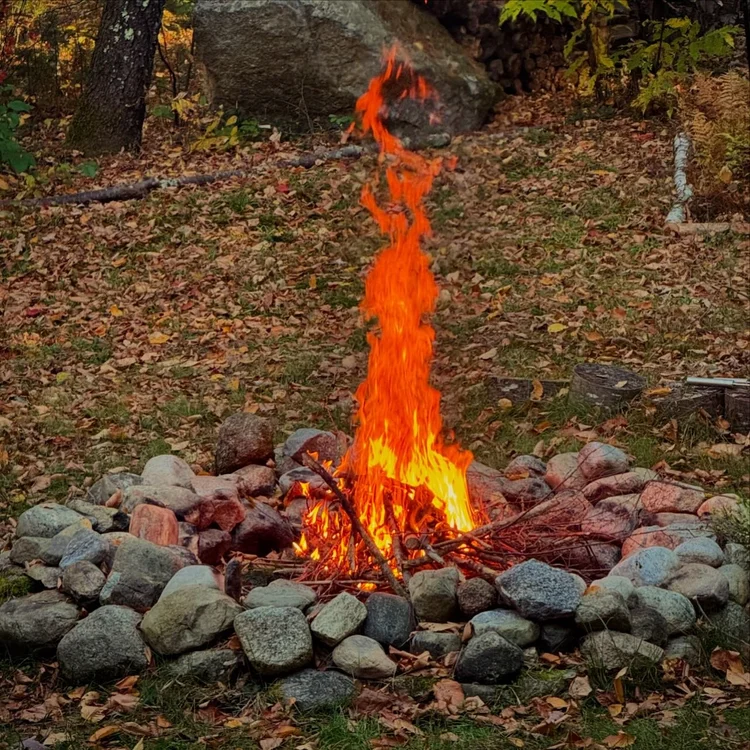 Bright flames rising from a rock-lined firepit in a backyard, with autumn leaves scattered around.
