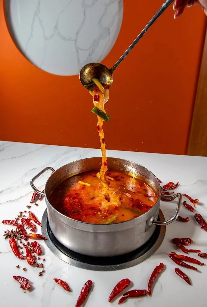 Spiced hot pot broth splashing in a steel pot beside fresh tomatoes and celery