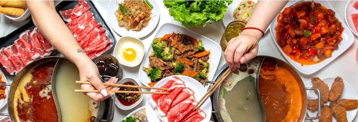 Overhead shot of hands sharing a split hot pot loaded with sliced meat and vegetables