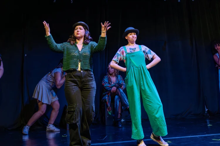 Two performers, one in green overalls, strike poses as the company plays behind them
