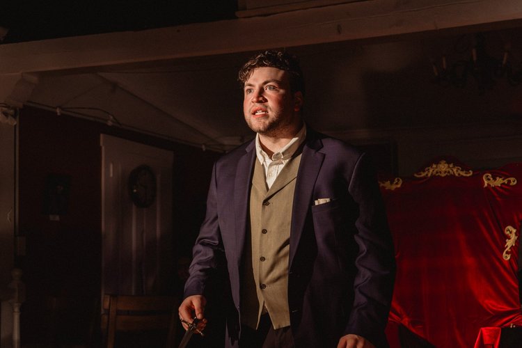 An actor in a three-piece suit stands in warm light against a red-curtained set