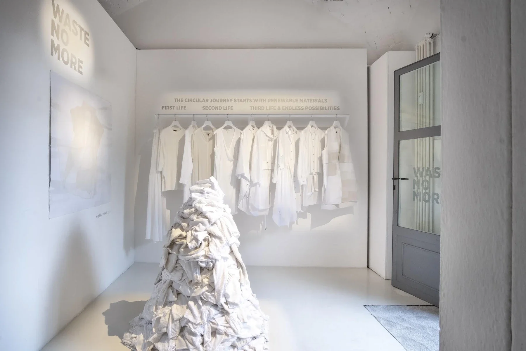 White garments hanging above a pile of clothing in the WASTE NO MORE exhibition in Milan.