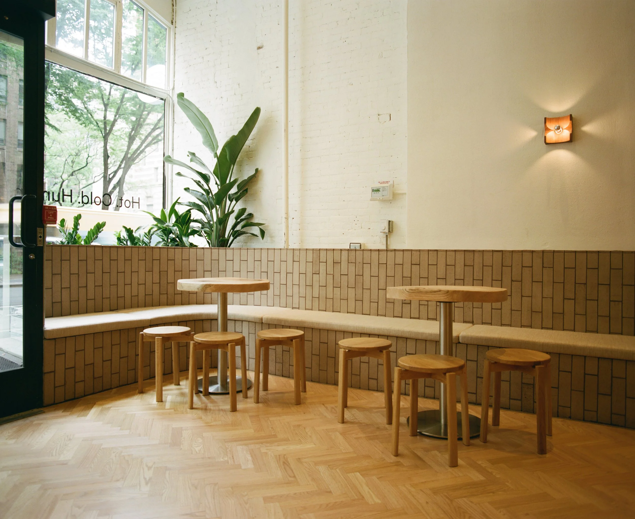 Wooden tables and stools beside a tiled banquette and tall window at Lore Bathing Club.