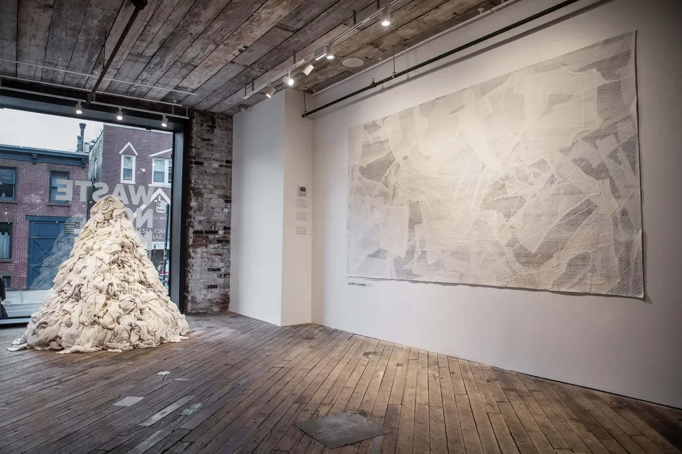 A tall pile of white clothing beside a textile artwork at Eileen Fisher’s WASTE NO MORE exhibition in the Brooklyn store.