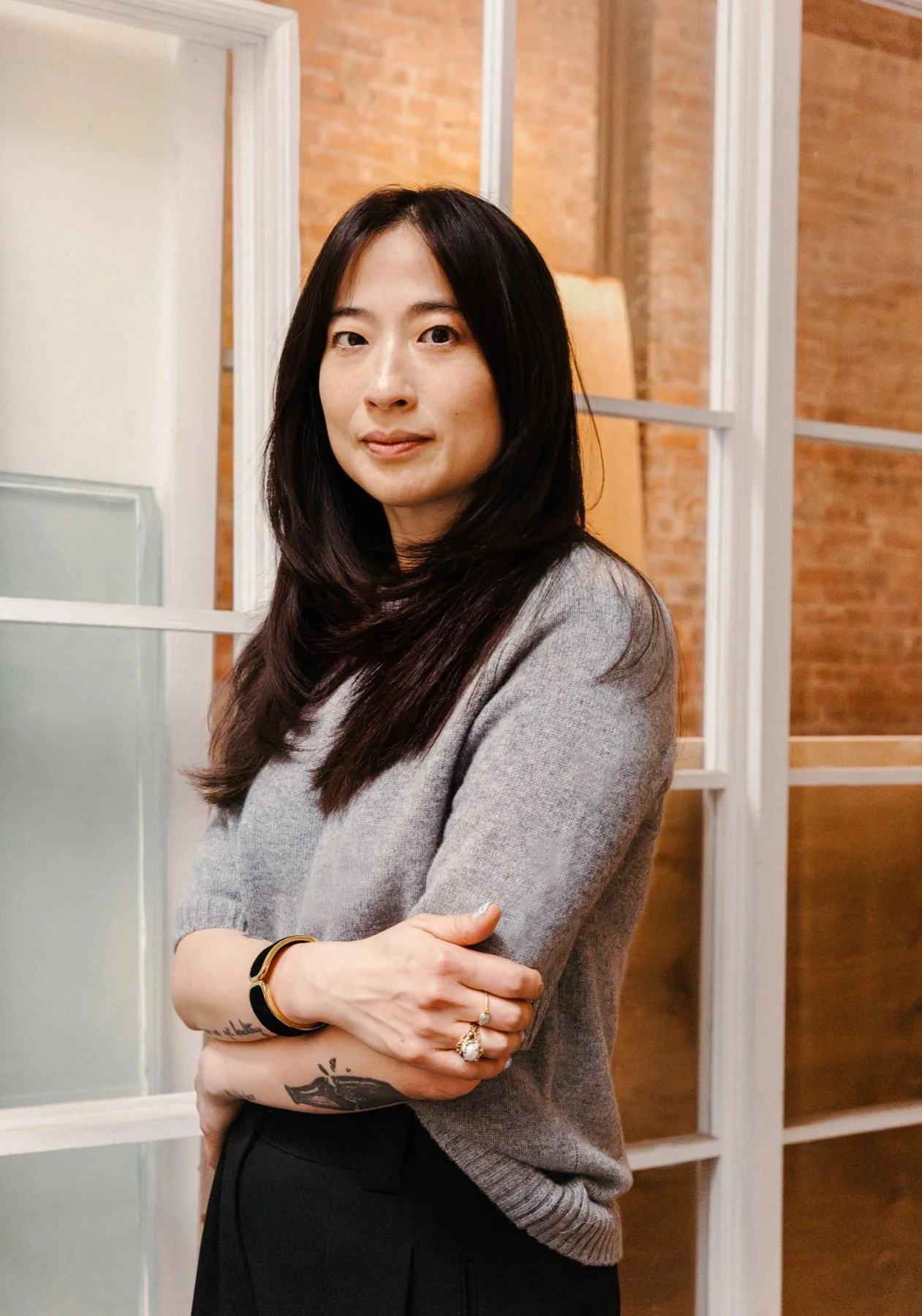 Jane Ivana Hong, photographed by Aly Kula, standing beside a window in a grey sweater.