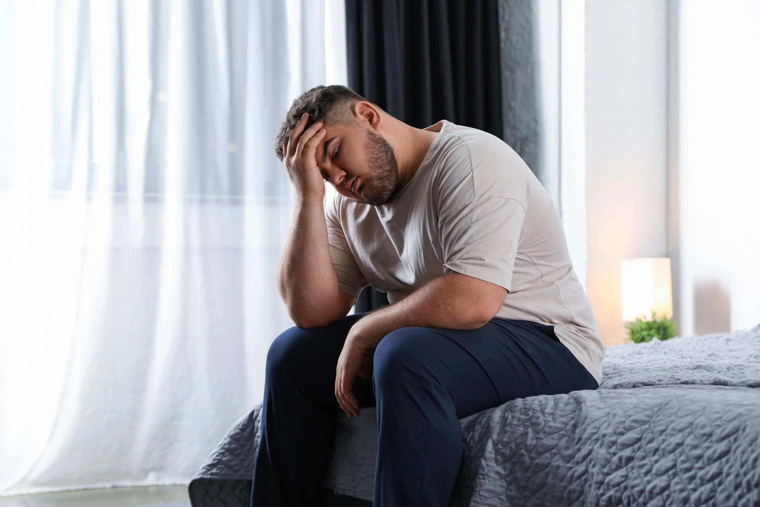 Tired man sitting on the edge of his bed — low energy and stubborn weight gain are common signs of low testosterone or thyroid issues in men