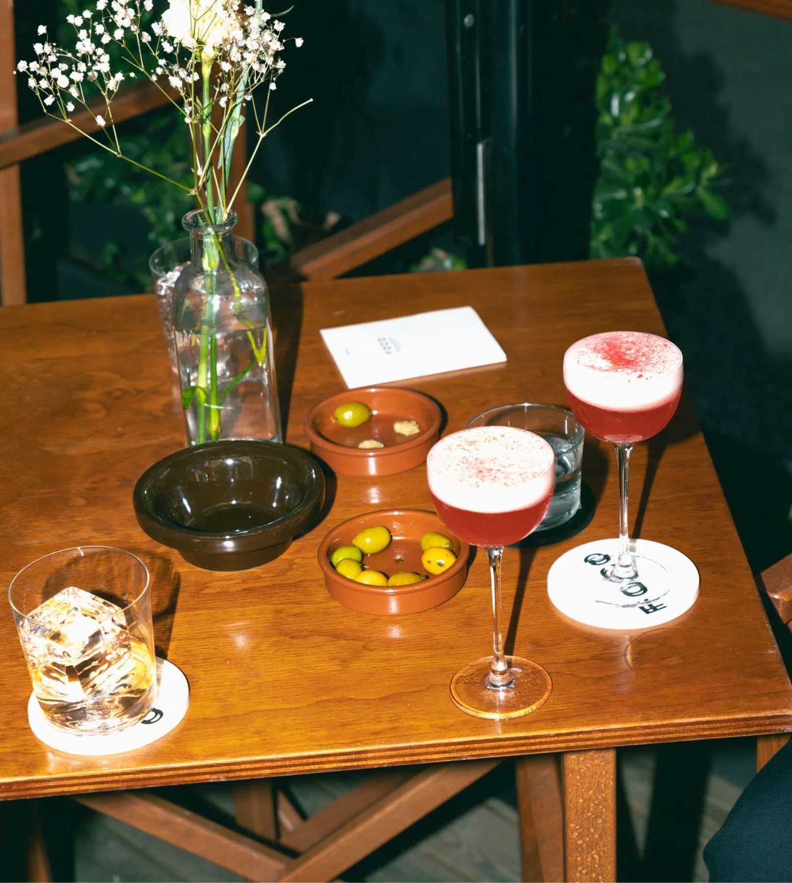Dos sours, un old fashioned i olives en una taula de la terrassa amb flors.
