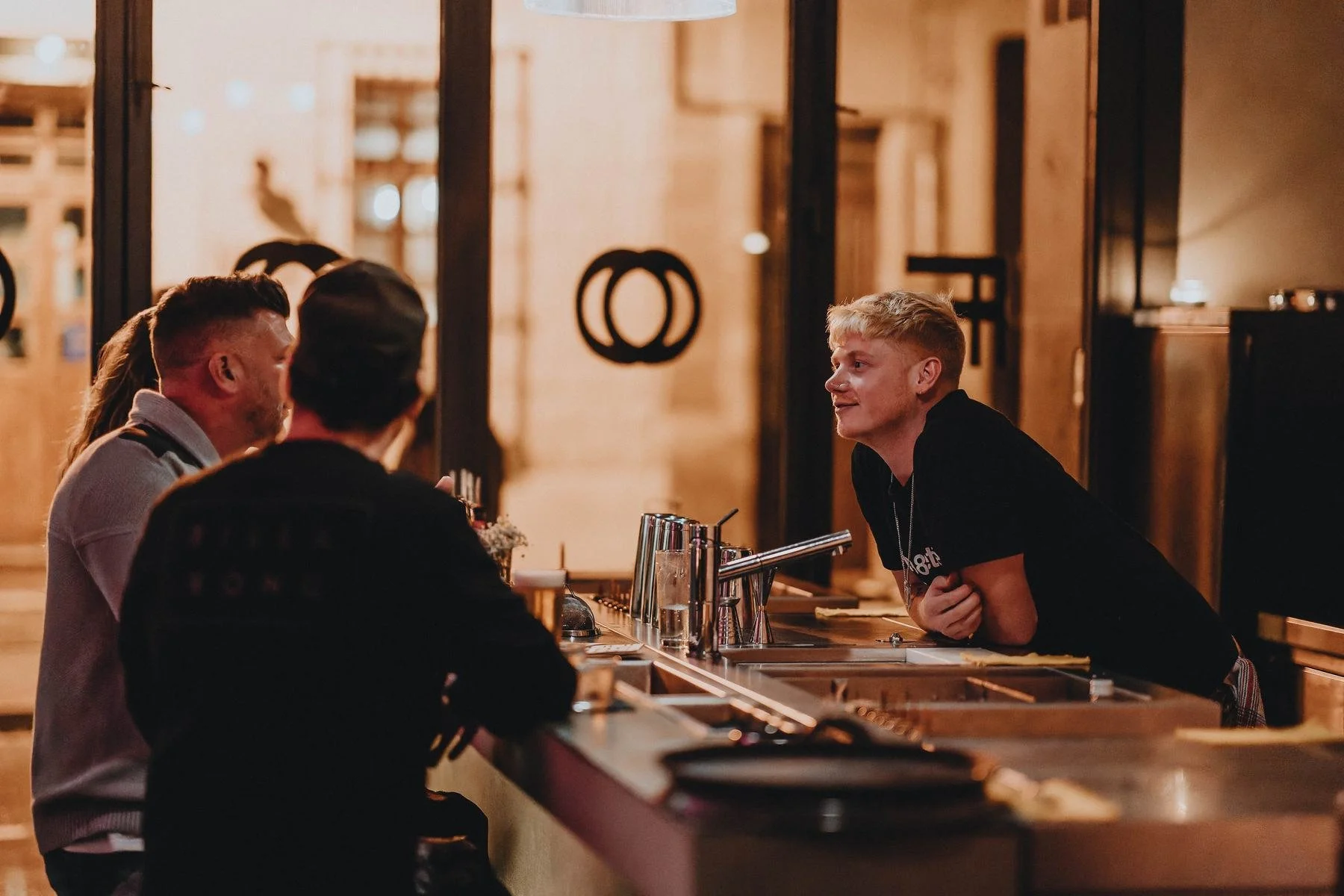 Un bartender hablando con dos clientes en la barra, de noche.