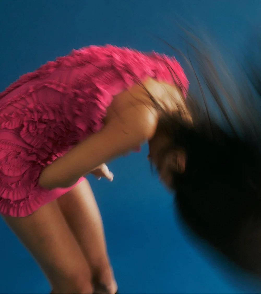 A model in a pink ruffled dress flipping her hair against a blue backdrop, from a Haus of Heir shoot