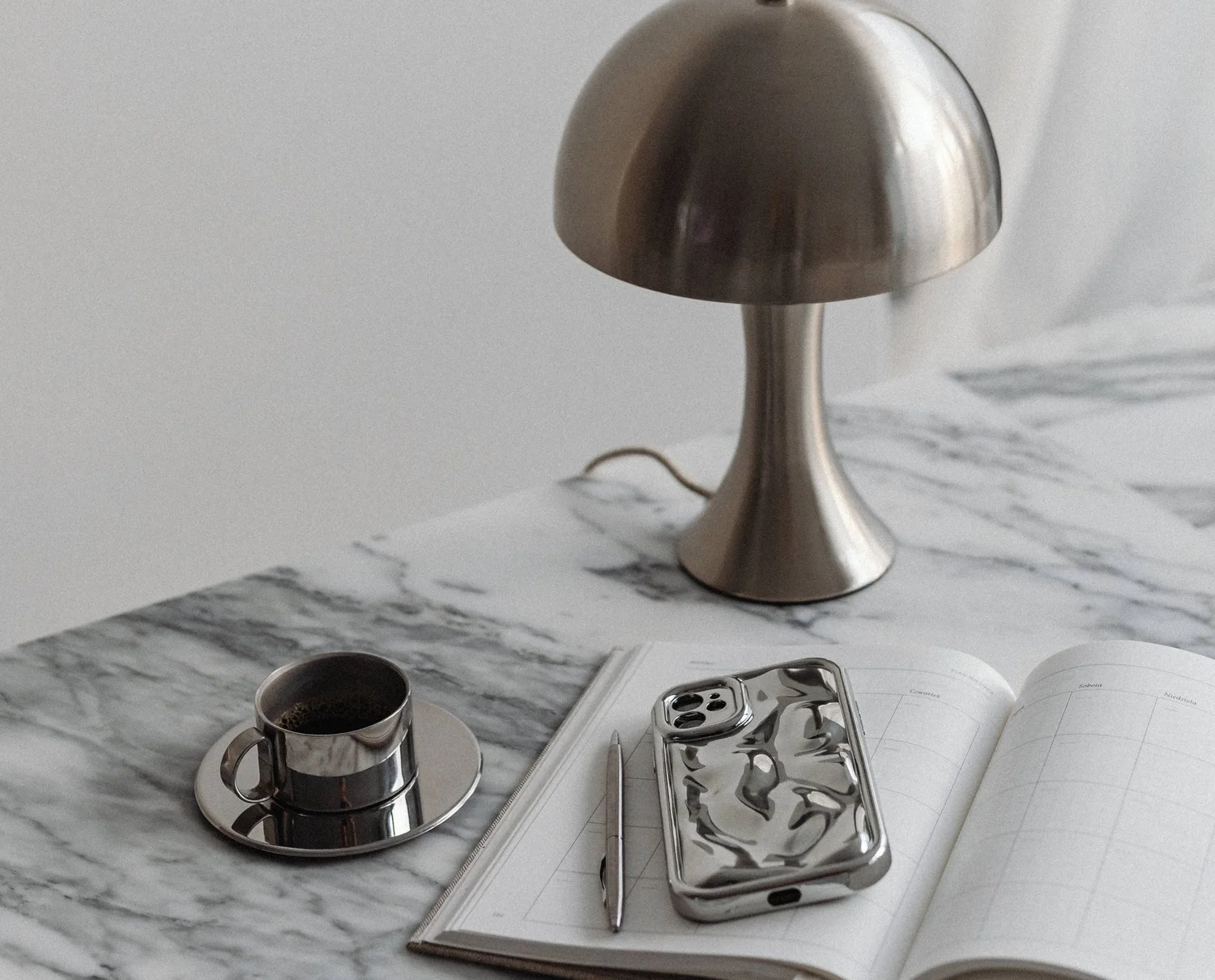 A chrome lamp, espresso cup and phone on a marble desk with a planner