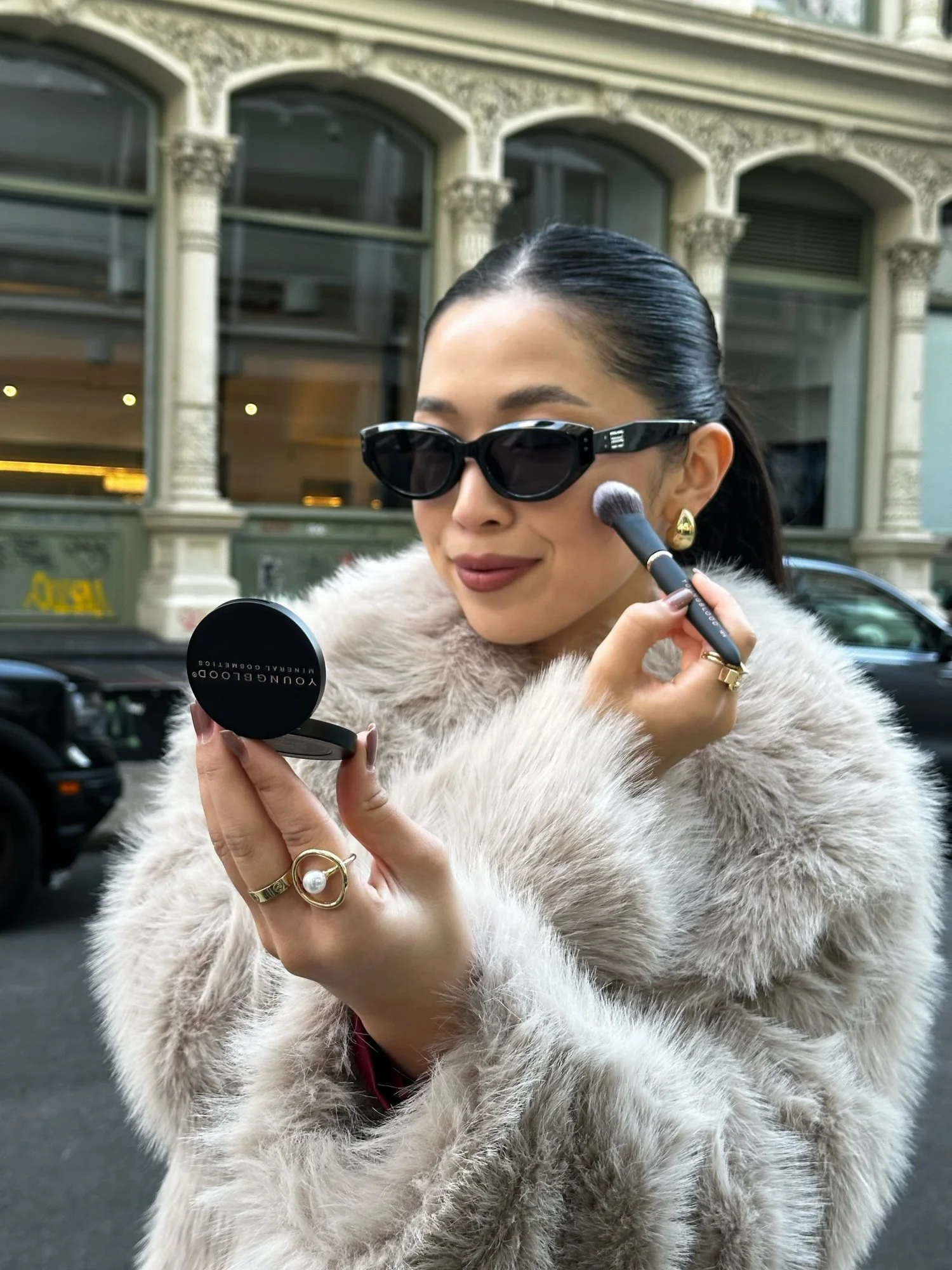A creator in sunglasses and a faux fur coat applying Youngblood powder on a city street