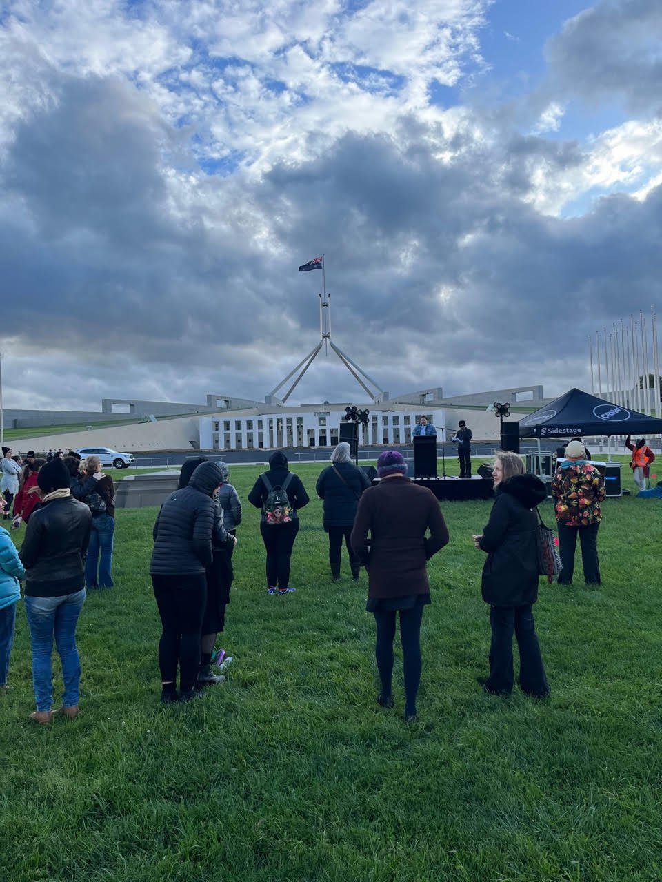 a group of people standing on grass facing a speaker at a podium in front of the Parliament House, Australia