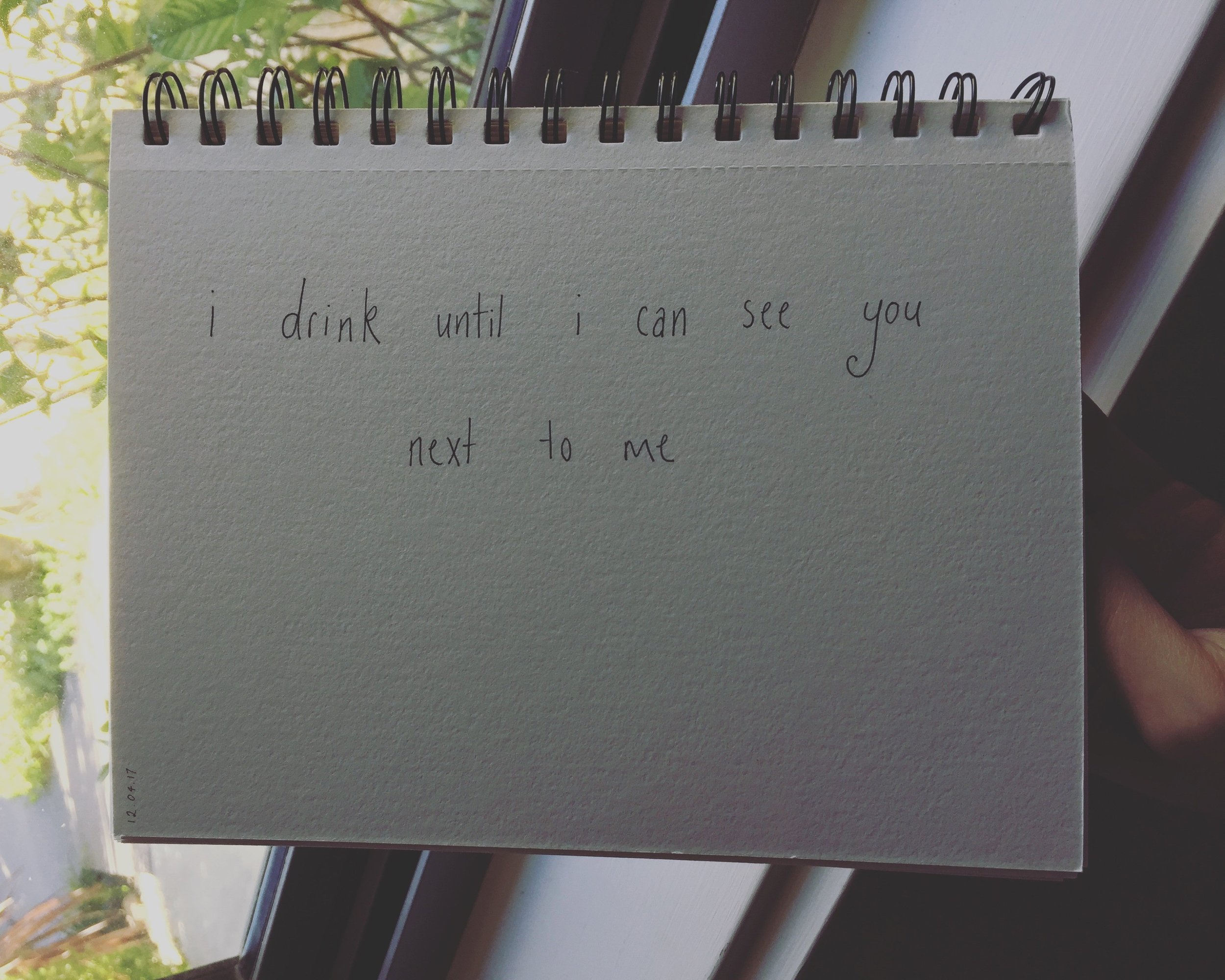 a hand holding a notebook that reads I drink until I can see you next to me on 