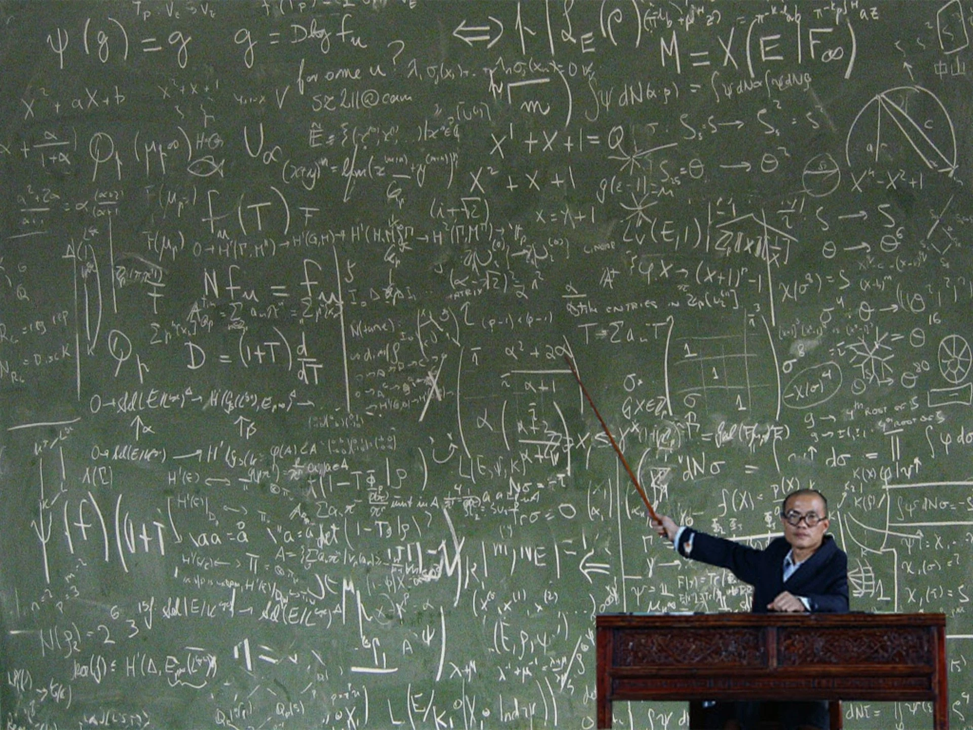A lecturer with a pointer at a chalkboard covered edge to edge in handwritten equations and diagrams