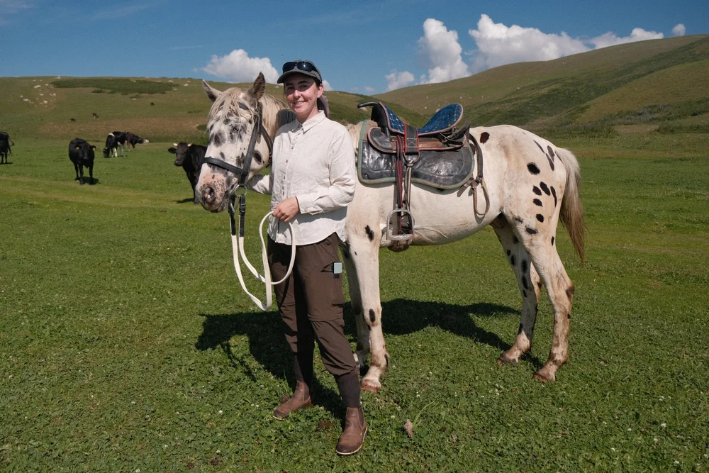 Kayla with a saddled spotted horse in a green mountain valley