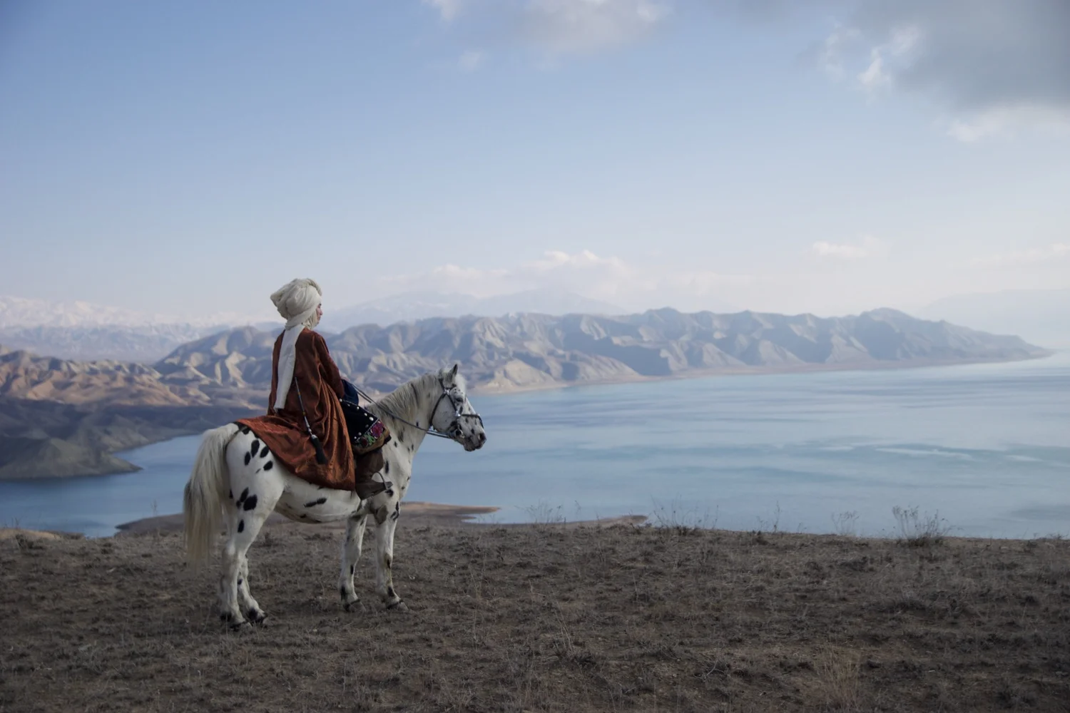 A Kyrgyz rider in traditional red dress on the spotted stallion Chaar Suluu, above a mountain lake