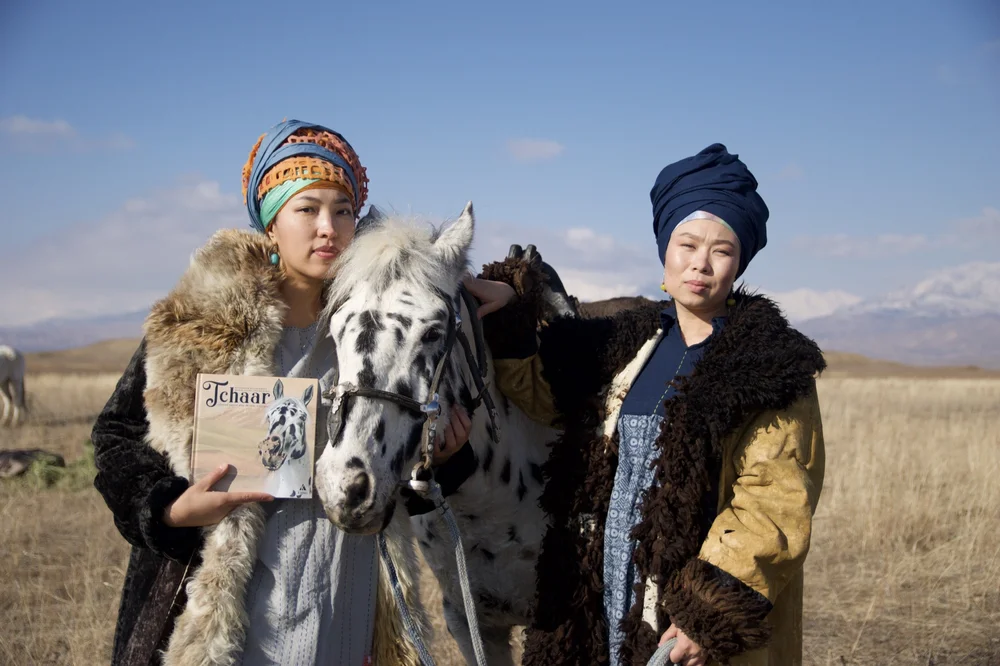 Two Kyrgyz women in traditional dress with Chaar Suluu, one holding Tchaar, the association's illustrated children's book of the Tolubai Synchy legend