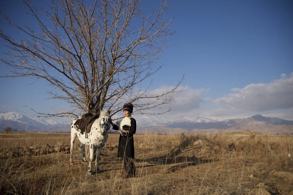 A Kyrgyz woman in traditional dress with the spotted stallion Chaar Suluu beneath a lone tree