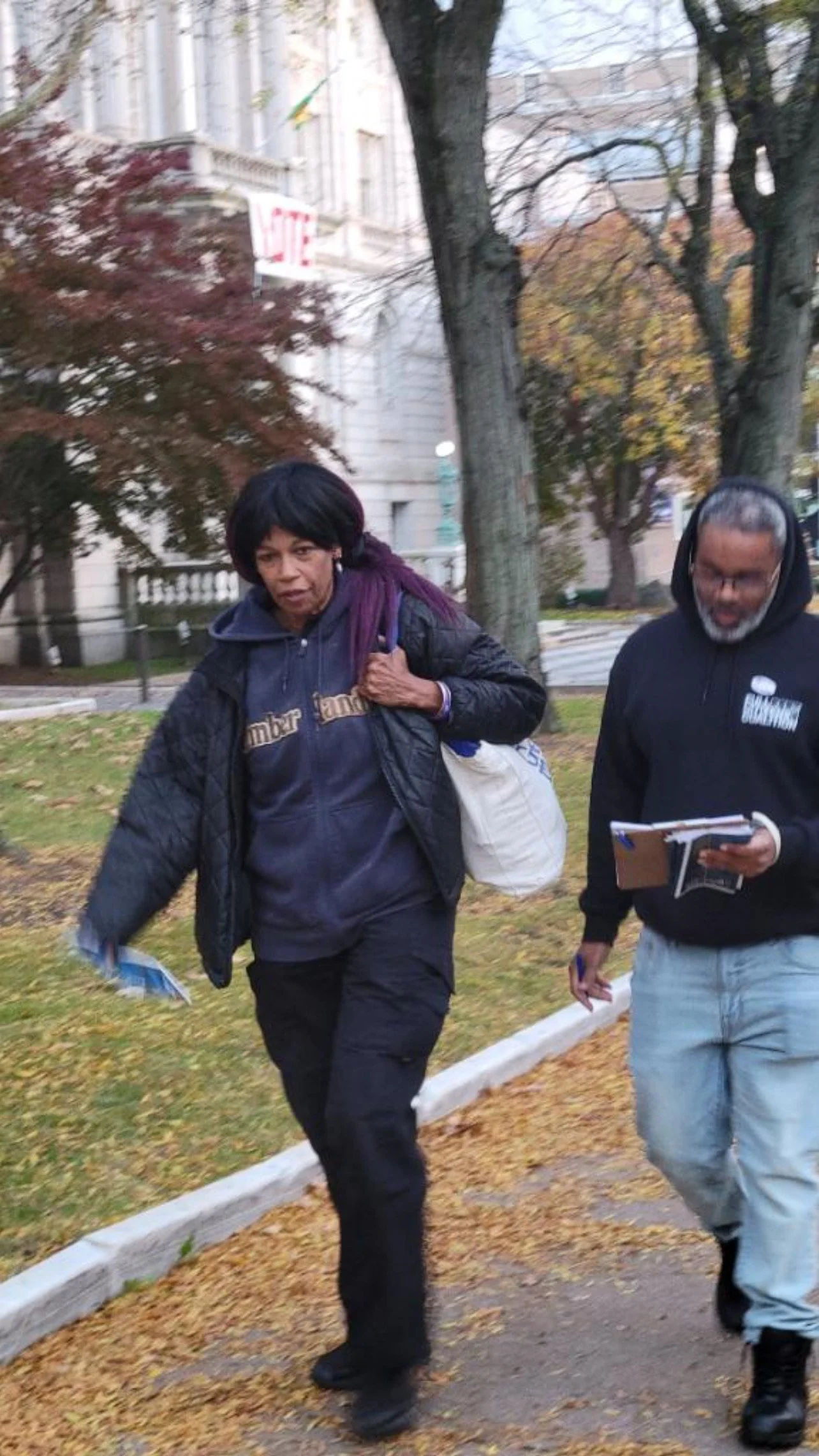 A canvasser with a clipboard walks with a woman past a building with a Vote sign